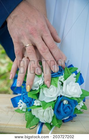 Hands of the newlyweds with gold rings on a bouquet of hand-made flowers with blue and white colors from the foamiran in the summer