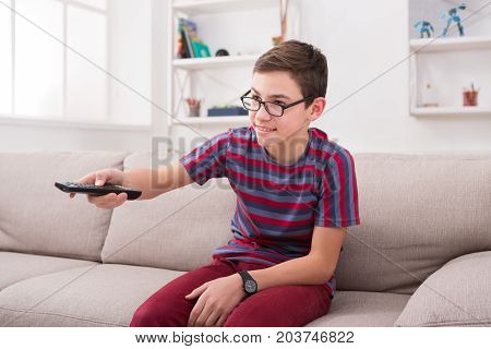 Teenager boy watching television, pointing with remote conroller while sitting on couch in living room at home