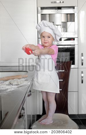 Little smiling baby girl baker in white cook hat and apron kneads a dough on tle kitchen.