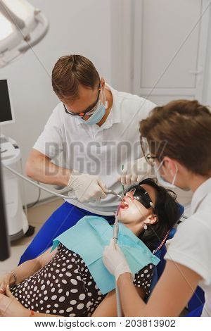 A Male Dentist With A Female Assistant Helps To Treat The Teeth Of A Woman Patient In A Clinic In Th