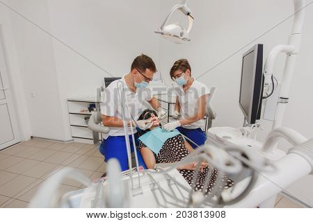 A Male Dentist With A Female Assistant Helps To Treat The Teeth Of A Woman Patient In A Clinic In Th