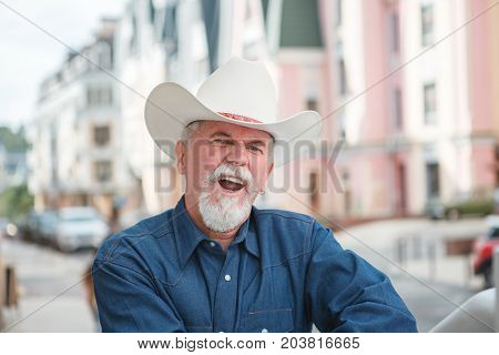 A mature cowboy in a hat jeans and a denim shirt looks at the camera. On open air
