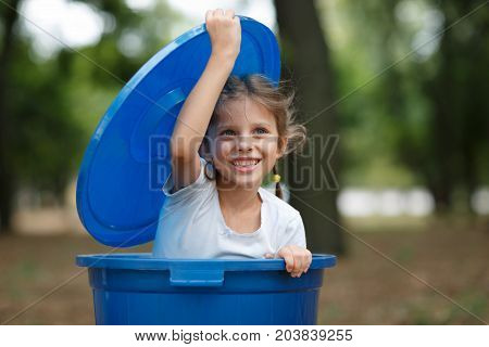 Smiling preschooler girl, playing with the trash bin lid, in the street, summer outdoor, save the planet, save the Earth concept. Day in park. Funny babies.