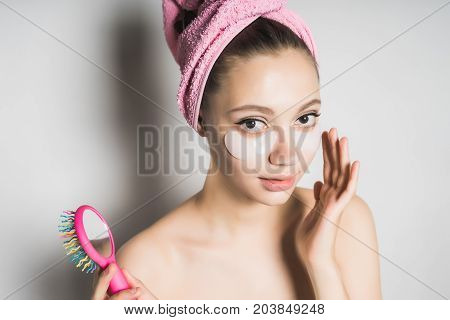 A young girl after bathing with a towel on her head takes care of the skin, holds in her hand a comb with a mirror, looks in surprise at the camera