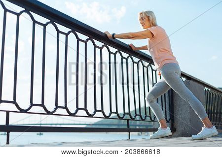 Improving suppleness. Athletic slender senior woman resting her hands on the balustrade and warming up before a morning run by doing stretching