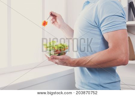 Man eat salad, healthy lunch in modern interior. Unrecognizable profile male torso in blue t-shirt, hand with fork, near window with vegetable salad in bowl, diet food concept.