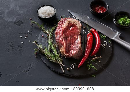 Raw rib eye steak with herbs and spices. Cooking ingredients for restaurant dish. Fresh meat with chilli and knife on plate on black background.