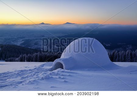 On the snowy lawn in the snowdrift there is an igloo covered by snow with the background of mountains forests fog and sunrise.