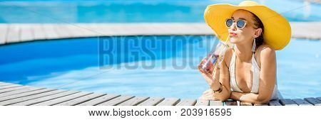 Young woman in swimsuit with big yellow sunhat relaxing with a bottle of fresh drink sitting on the poolside outdoors. Panoramic cropped image