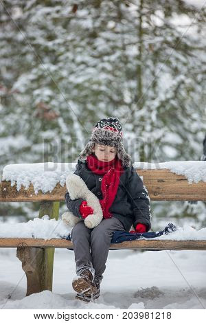 Two Children, Boy Brothers, Sitting On A Bench In Park, Wintertime While Snowing