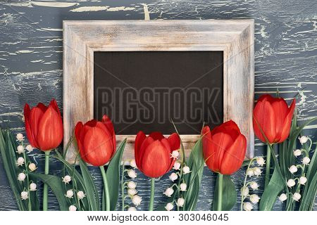 Blackboard And Bunch Of Red Tulips And Lily Of The Valley Flowers On Rustic Background. Space For Yo