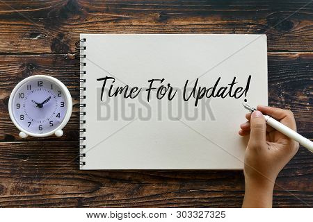 Top View Of Clock And Hand Holding Pen Writing Time For Update! On Notebook On Wooden Background.