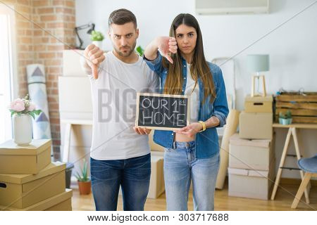 Young beautiful couple holding blackboard with our first home text at new house with angry face, negative sign showing dislike with thumbs down, rejection concept