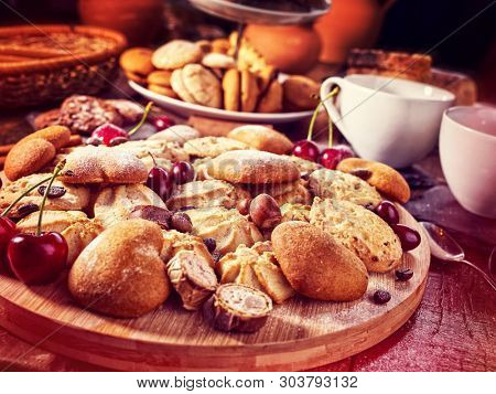 Oatmeal cookies and sand chocolate cake with cherry on cutting board on wooden table in rustic style served with cups and dessert spoons. Sun flare morning sunrays from window. Dark environment.