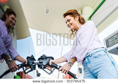 Low Angle View Of Happy Woman Holding Fuel Pump While Refueling Car With Benzine