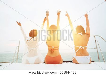 Rear View Of Three Cheerful And Happy Young Women In Bikini Sitting On Sailing Yacht Boat And Admiri