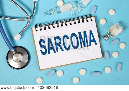 Sarcoma, Text On White Notepad Over Blue Background. Blue Stethoscope. Pills. Ampoules