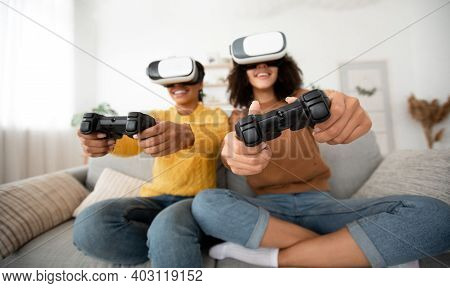 Females Roommates Have Fun On Couch In Cozy Interior With Video Games. Enthusiastic Smiling Teen Afr