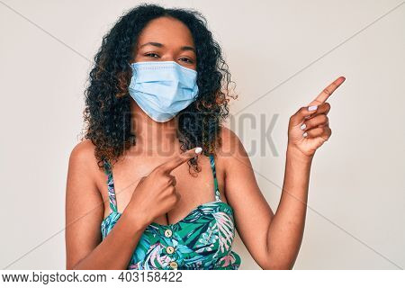 Young african american woman wearing medical mask smiling and looking at the camera pointing with two hands and fingers to the side. 