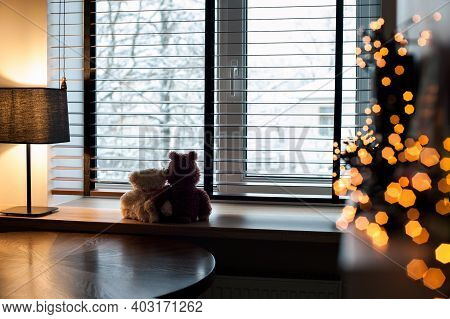 Cozy home evening-two loving embracing teddy bear toys sitting together on wooden windowsill with burning lamp and garland light decor. Living room interior. Filtered horizontal indoor image