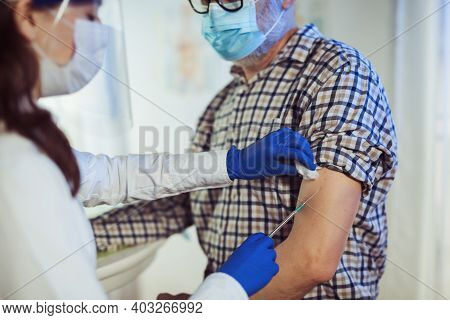 Young Woman Nurse With Surgical Mask And Face Shild Giving Injection To Senior Man. Covid-19 Or Coro