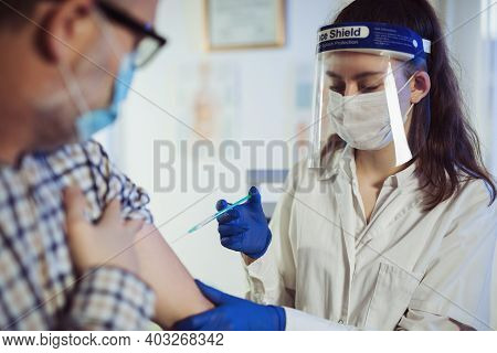 Young Woman Nurse With Surgical Mask And Face Shild Giving Injection To Senior Man. Covid-19 Or Coro