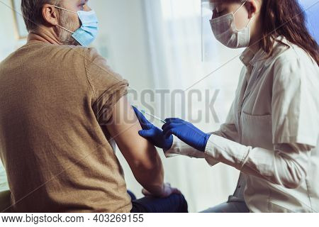 Young Woman Nurse With Surgical Mask And Face Shild Giving Injection To Senior Man. Covid-19 Or Coro