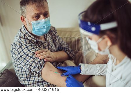 Young Woman Nurse With Surgical Mask And Face Shild Giving Injection To Senior Man At Home Or In A N