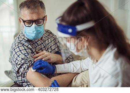 Young Woman Nurse With Surgical Mask And Face Shild Giving Injection To Senior Man At Home Or In A N