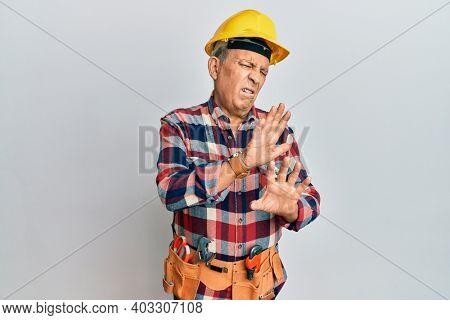 Senior hispanic man wearing handyman uniform disgusted expression, displeased and fearful doing disgust face because aversion reaction. with hands raised 