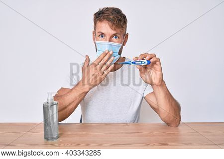 Young caucasian man wearing medical mask, hand sanitizer gel and thermometer sitting on the table covering mouth with hand, shocked and afraid for mistake. surprised expression 