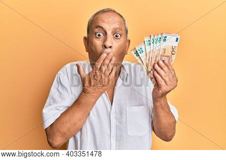 Handsome mature man holding bunch of 50 euro banknotes covering mouth with hand, shocked and afraid for mistake. surprised expression 