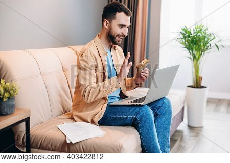 Man Speaking By Videocall, Eating Sandwich And Sitting On Sofa In His Cozy Living Room. Man Communic