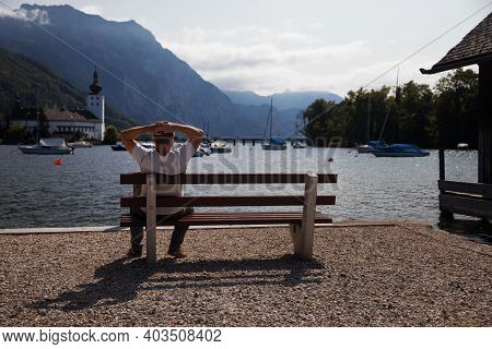 The Middle Aged Man Relaxing By Lake, Watching At Water And Mountains. Gmunden, Austria.