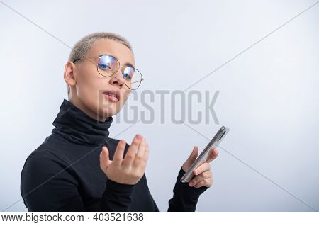 Young Woman With Short Blonde Haircut In A Black Turtleneck And Glasses Uses A Smartphone On A White