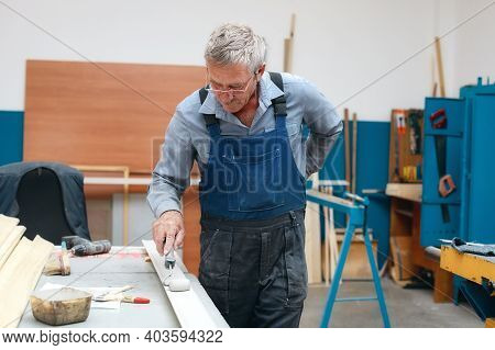 An Elderly Cabinetmaker In Overalls And Glasses Paints A Wooden Board With A Roller On A Workbench I
