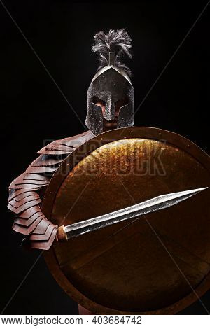 Gladiator With Sword And Armor On A Black Background. A Warrior In Gladiatorial Armor And A Helmet H