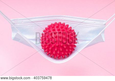 A Red Ball With Spikes Coronavirus Swinging In A Medical Mask Like In A Hammock Or Swing.