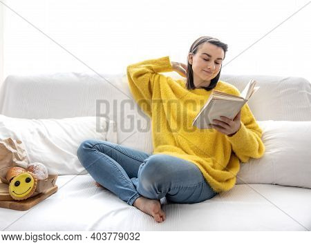 Stylish Girl In A Yellow Sweater Is Resting At Home On The Sofa With A Book.