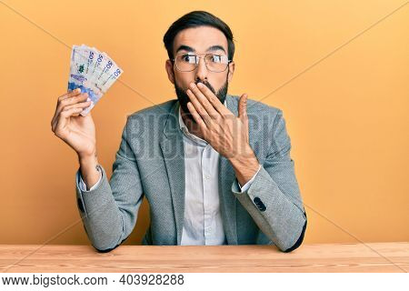 Young hispanic man holding colombian pesos banknotes sitting on the table covering mouth with hand, shocked and afraid for mistake. surprised expression 