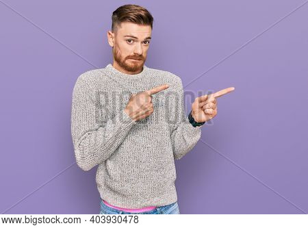 Young redhead man wearing casual winter sweater pointing aside worried and nervous with both hands, concerned and surprised expression 