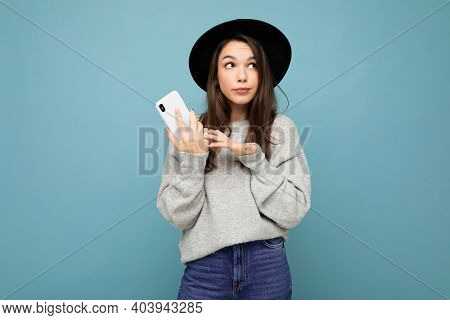 Beautiful Young Brunette Woman Thinking Wearing Black Hat And Grey Sweater Holding Smartphone Lookin