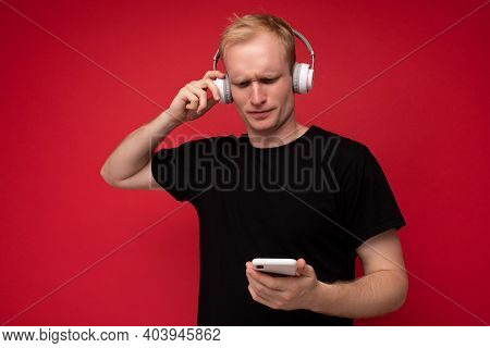 Upset Thoughtful Handsome Blonde Young Man Wearing Black T-shirt And White Headphones Standing Isola