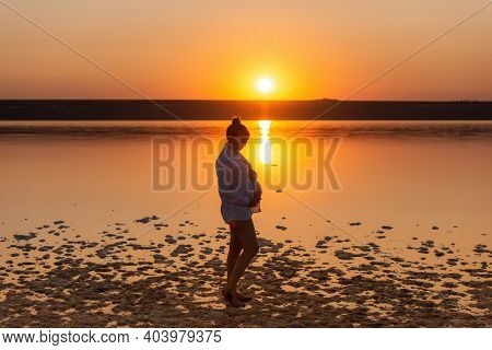 Silhouette Of A Pregnant Woman On The Beach At Sunset. A Pregnant Girl Against The Background Of The