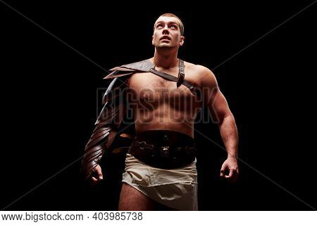 Gladiator With Sword And Armor On A Black Background. A Warrior In Gladiatorial Armor Looks Up At A 