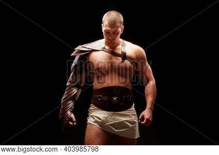 Gladiator With Sword And Armor On A Black Background. A Warrior In Gladiatorial Armor Looks Dawn At 