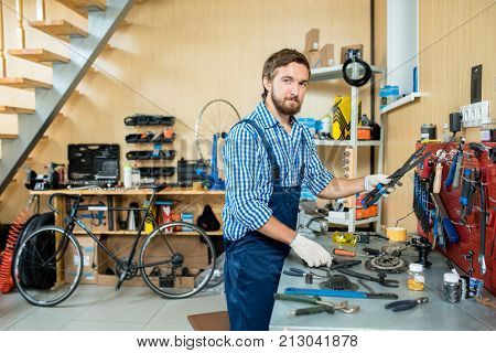 Young man in uniform and gloves looking at camera by his workplace in repair workshop