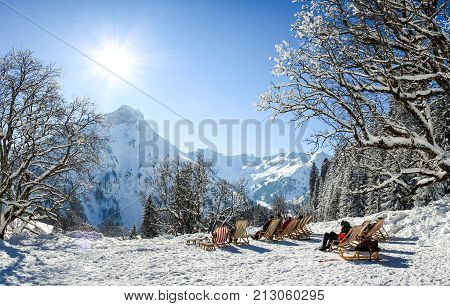 Group of people sitting with deck chairs in sunny winter mountains. Sunbathing in snow. Germany, Bavaria, Allgau, Schwarzenberghuette.