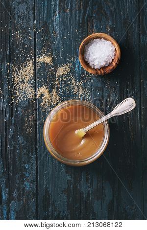 Glass jar of homemade salted caramel sauce with spoon, brown sugar and bowl of salt. Over old dark blue wooden background. Top view with space. Toned image