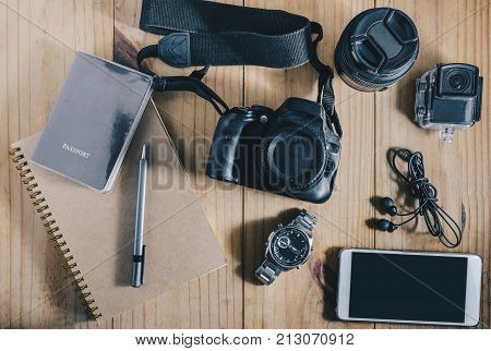 Top view of travel object : passport and gray pencil on brown notebook. and white mobilephone earphone black camera and two lens and actioncam on wooden table